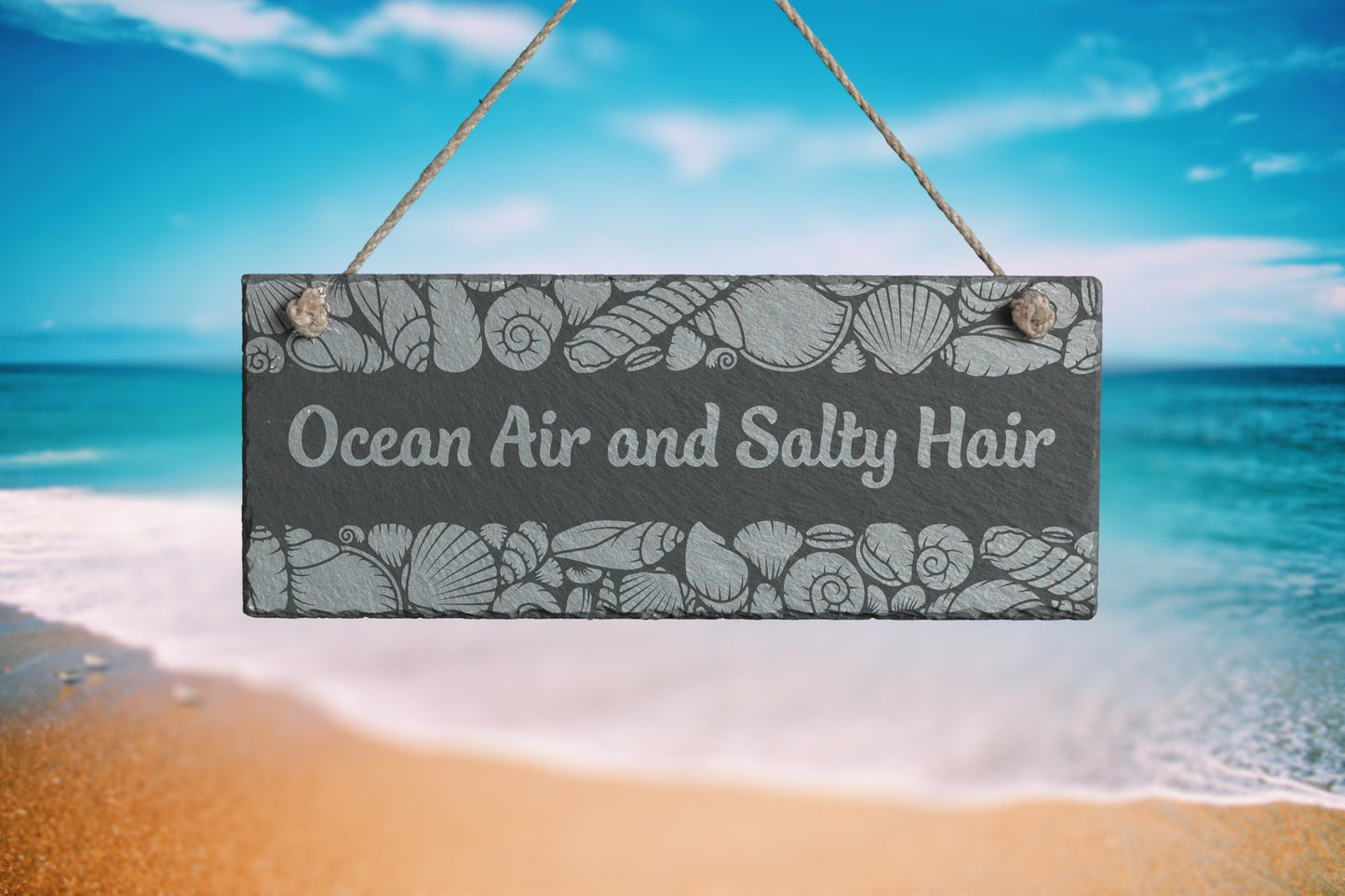 Customizable slate sign with engraved sea shell border and the text 'Ocean Air and Salty Hair', hanging in a beach setting.