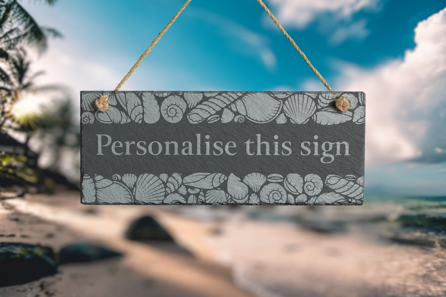 Decorative slate sign with seashell pattern and text 'Personalise this sign' against a beach backdrop