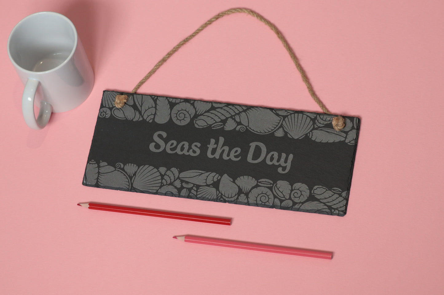 Flat-lay of a customizable slate sign with sea shells engraving and 'Seas the Day' text, next to a white mug and two coloured pencils on a light pink background.