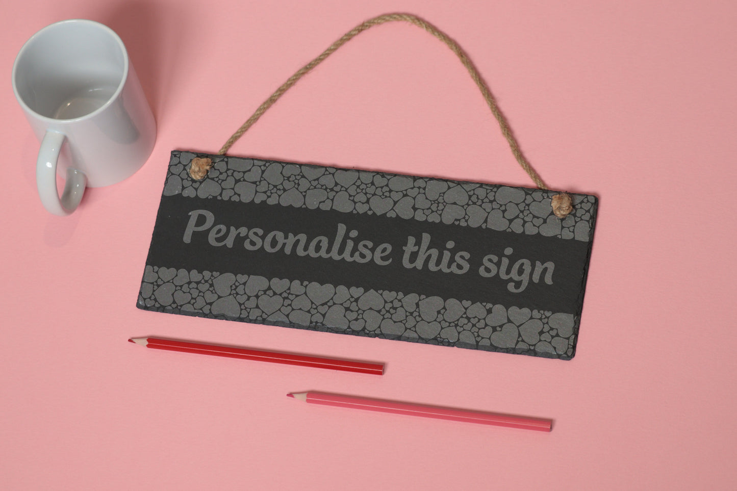 Flat-lay of a customizable slate sign with heart engraving and 'Personalise this sign' text, next to a white mug and two coloured pencils on a light pink background.