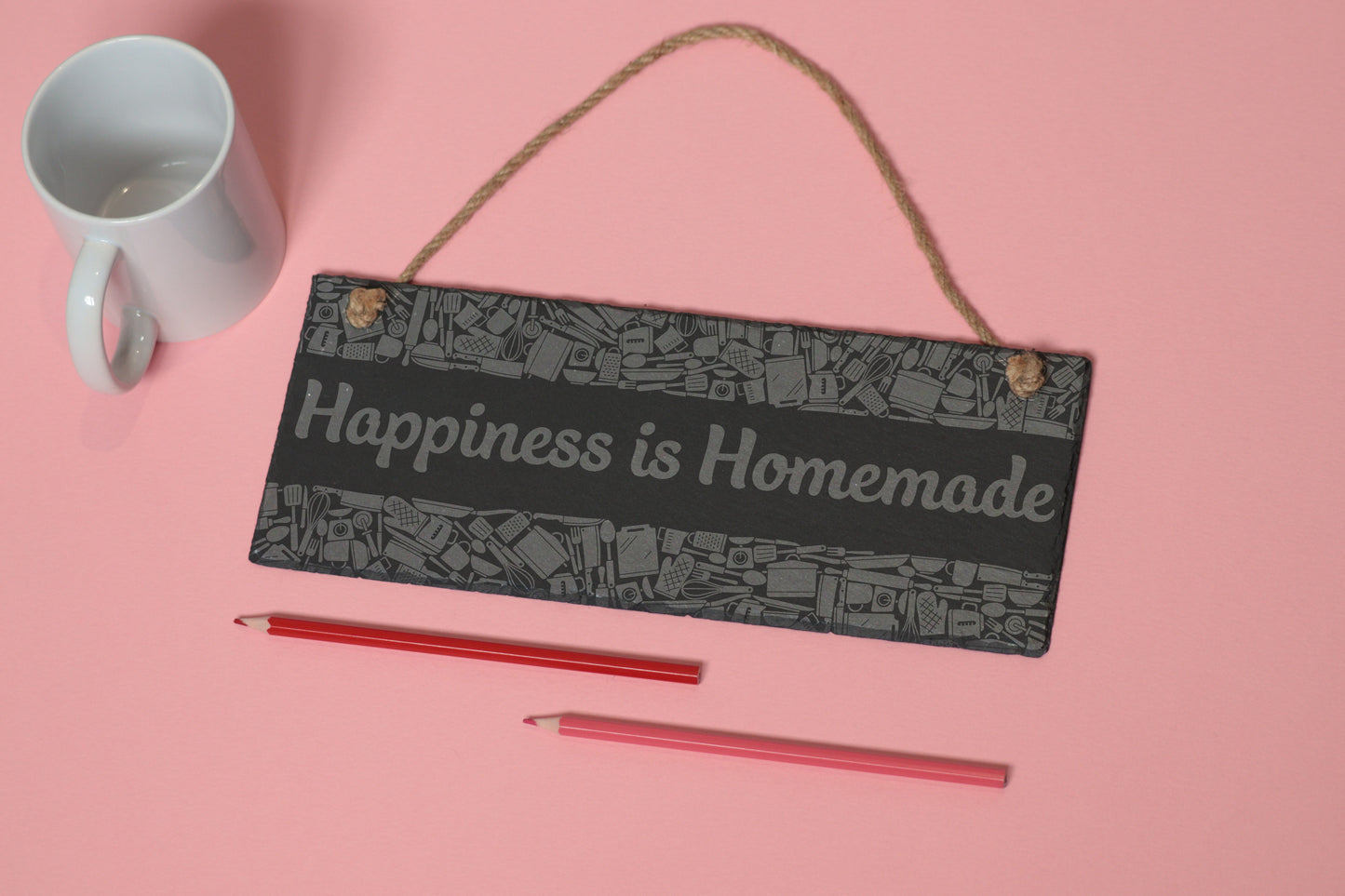 Flat-lay of a customizable slate sign with kitchen utensils engraving and 'Happiness is Homemade' text, next to a white mug and two coloured pencils on a light pink background.