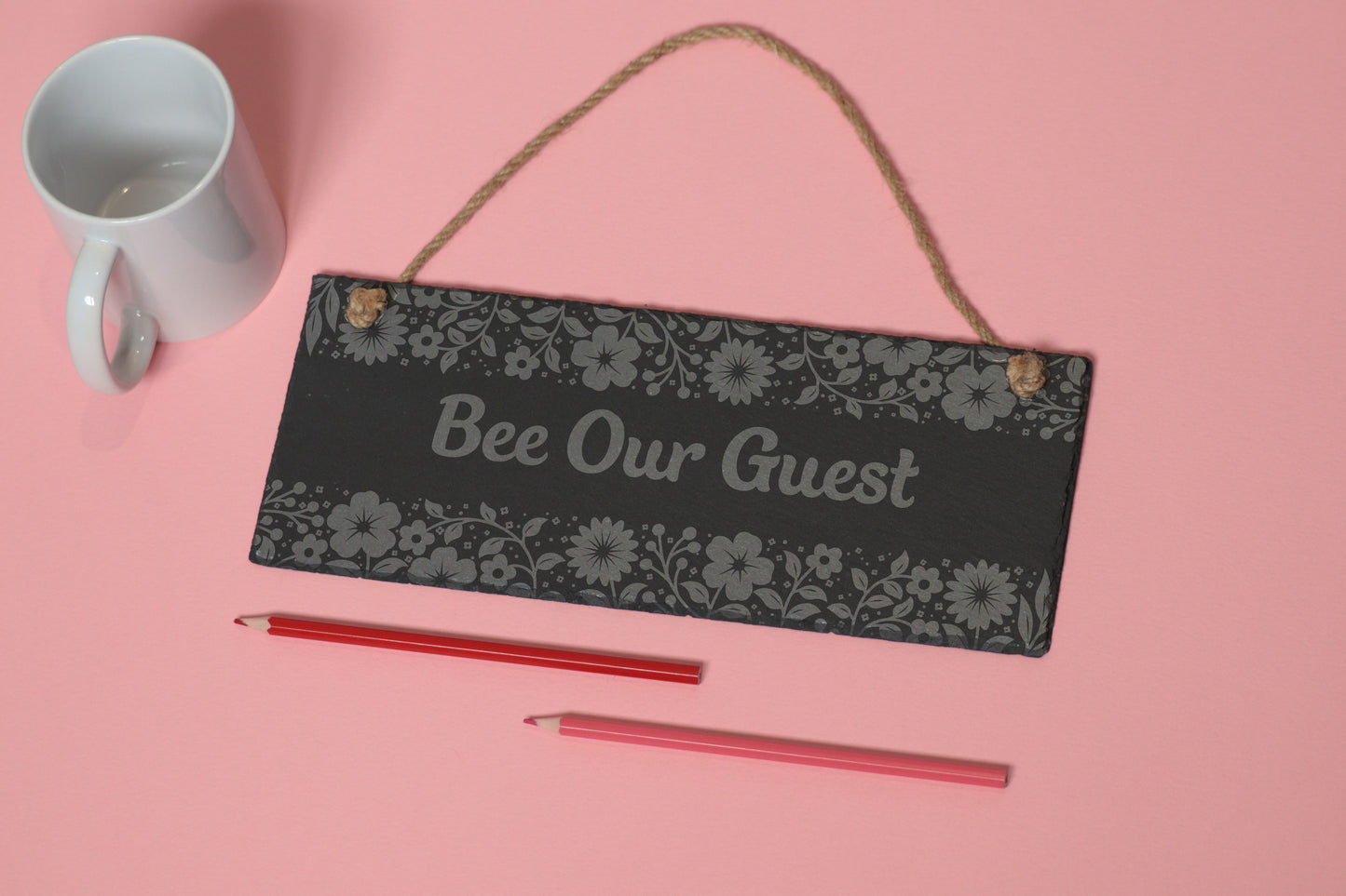 Flat-lay of a customizable slate sign with floral engraving and 'Bee Our Guest' text, next to a white mug and two coloured pencils on a light pink background.