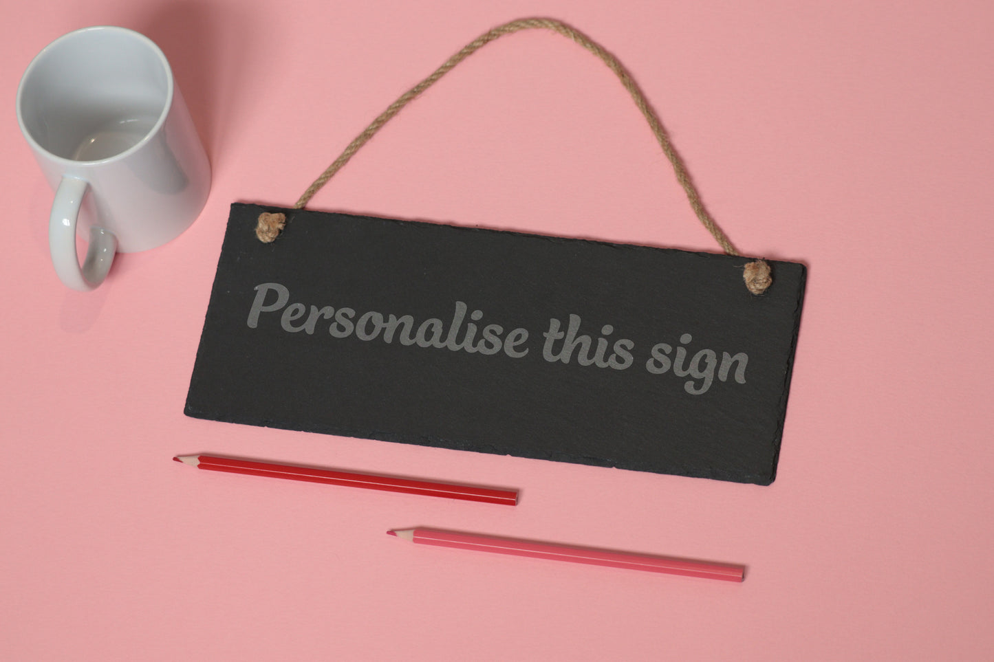 Flat-lay of a customizable slate sign with 'Personalise this sign' text, next to a white mug and two coloured pencils on a light pink background.