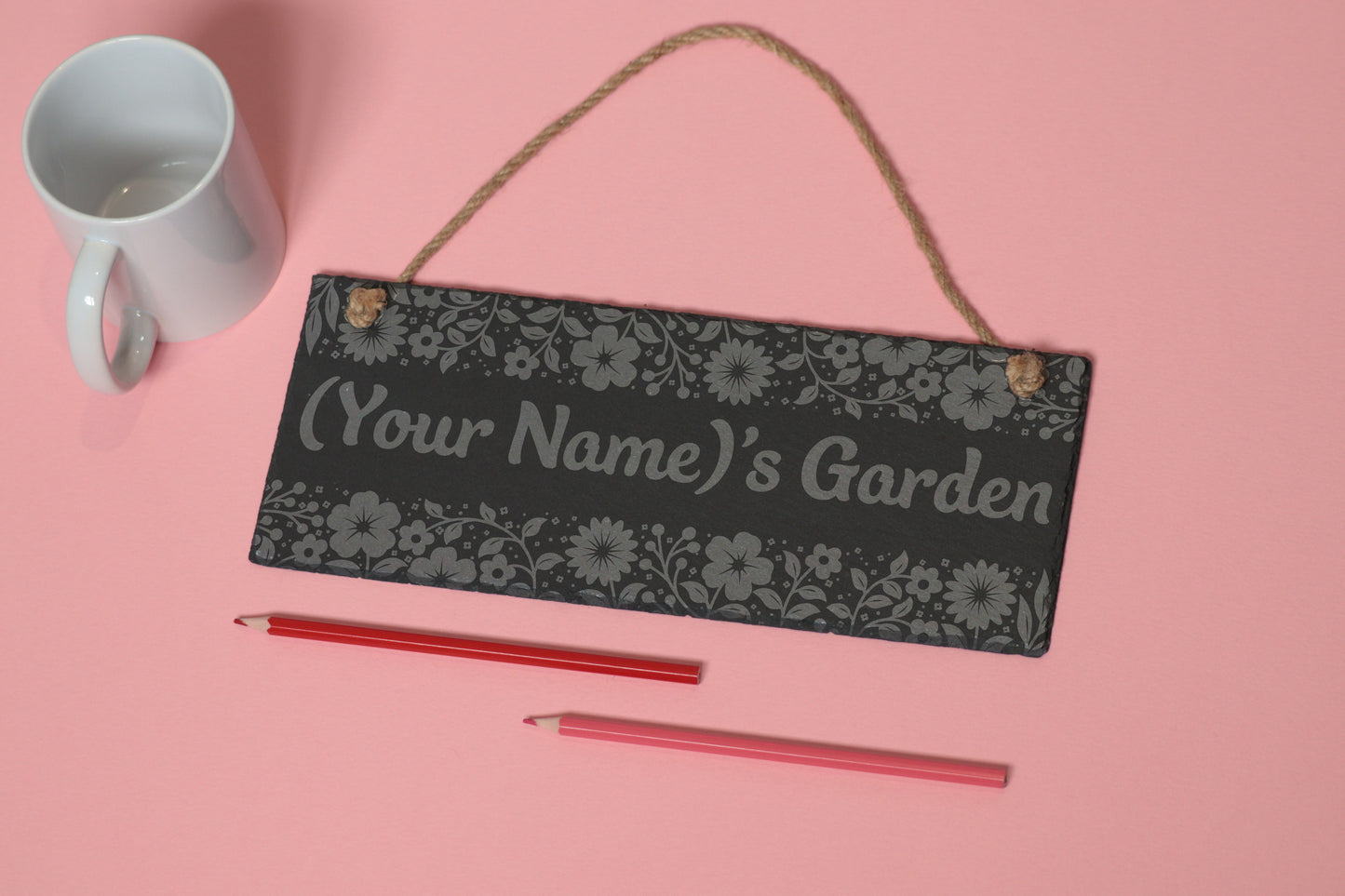 Flat-lay of a customizable slate sign with floral engraving and '(Your Name)'s Garden' text, next to a white mug and two coloured pencils on a light pink background.