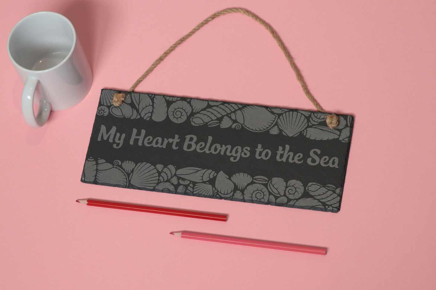 Flat-lay of a customizable slate sign with sea shells engraving and 'My Heart Belongs to the Sea' text, next to a white mug and two coloured pencils on a light pink background.
