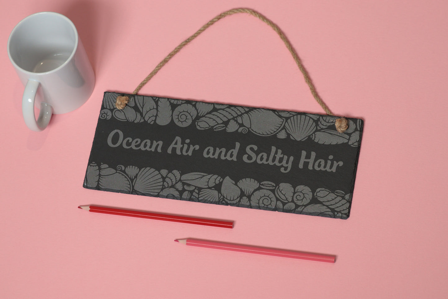 Flat-lay of a customizable slate sign with sea shells engraving and 'Ocean Air and Salty Hair' text, next to a white mug and two coloured pencils on a light pink background.
