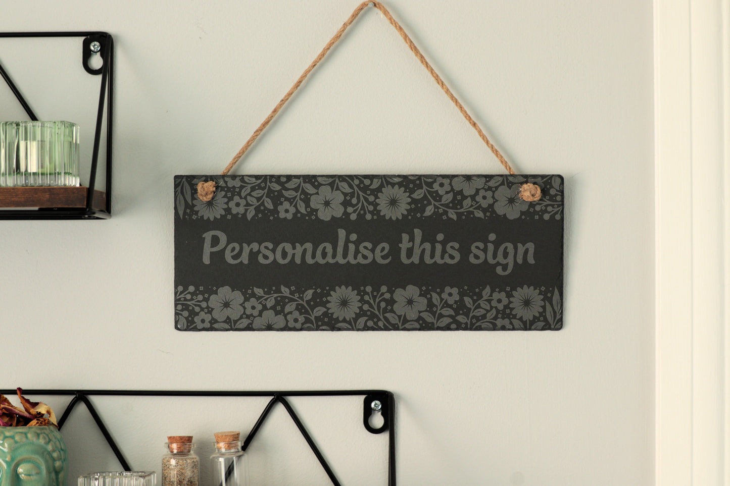 Customizable slate sign with floral border and the text 'Personalise this sign', hanging on a wall near shelves.