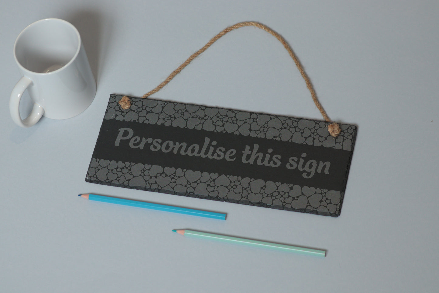 Flat-lay of a customizable slate sign with heart engraving and 'Personalise this sign' text, next to a white mug and two coloured pencils on a light blue background.