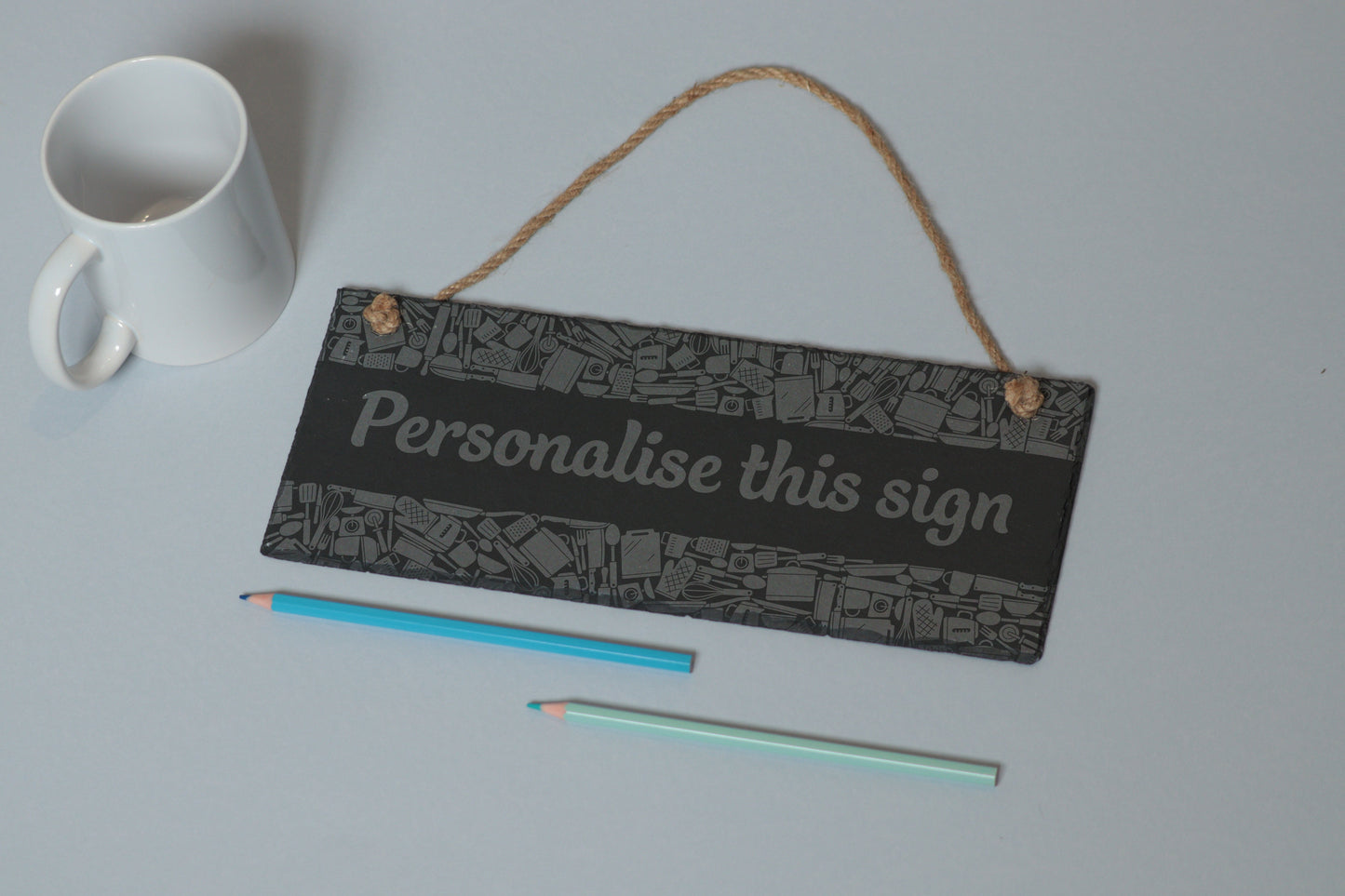 Flat-lay of a customizable slate sign with kitchen utensil engraving and 'Personalise this sign' text, next to a white mug and two coloured pencils on a light blue background.