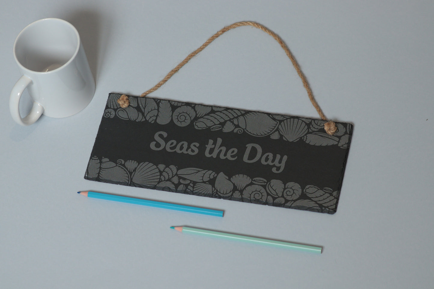 Flat-lay of a customizable slate sign with sea shell engraving and 'Seas the Day' text, next to a white mug and two coloured pencils on a light blue background.
