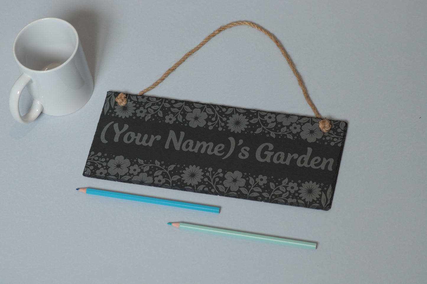 Flat-lay of a customizable slate sign with floral utensil engraving and '(Your Name)'s Garden' text, next to a white mug and two coloured pencils on a light blue background.
