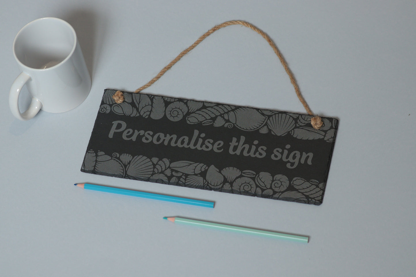 Flat-lay of a customizable slate sign with sea shell engraving and 'Personalise this sign' text, next to a white mug and two coloured pencils on a light blue background.