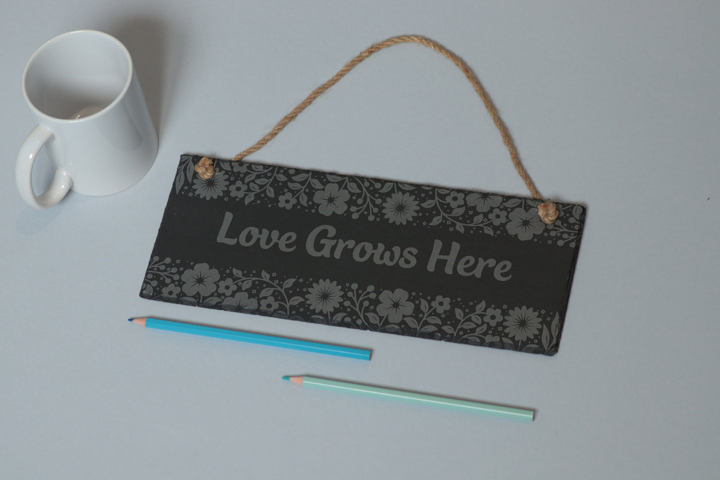 Flat-lay of a customizable slate sign with floral engraving and 'Love Grows Here' text, next to a white mug and two coloured pencils on a light blue background.