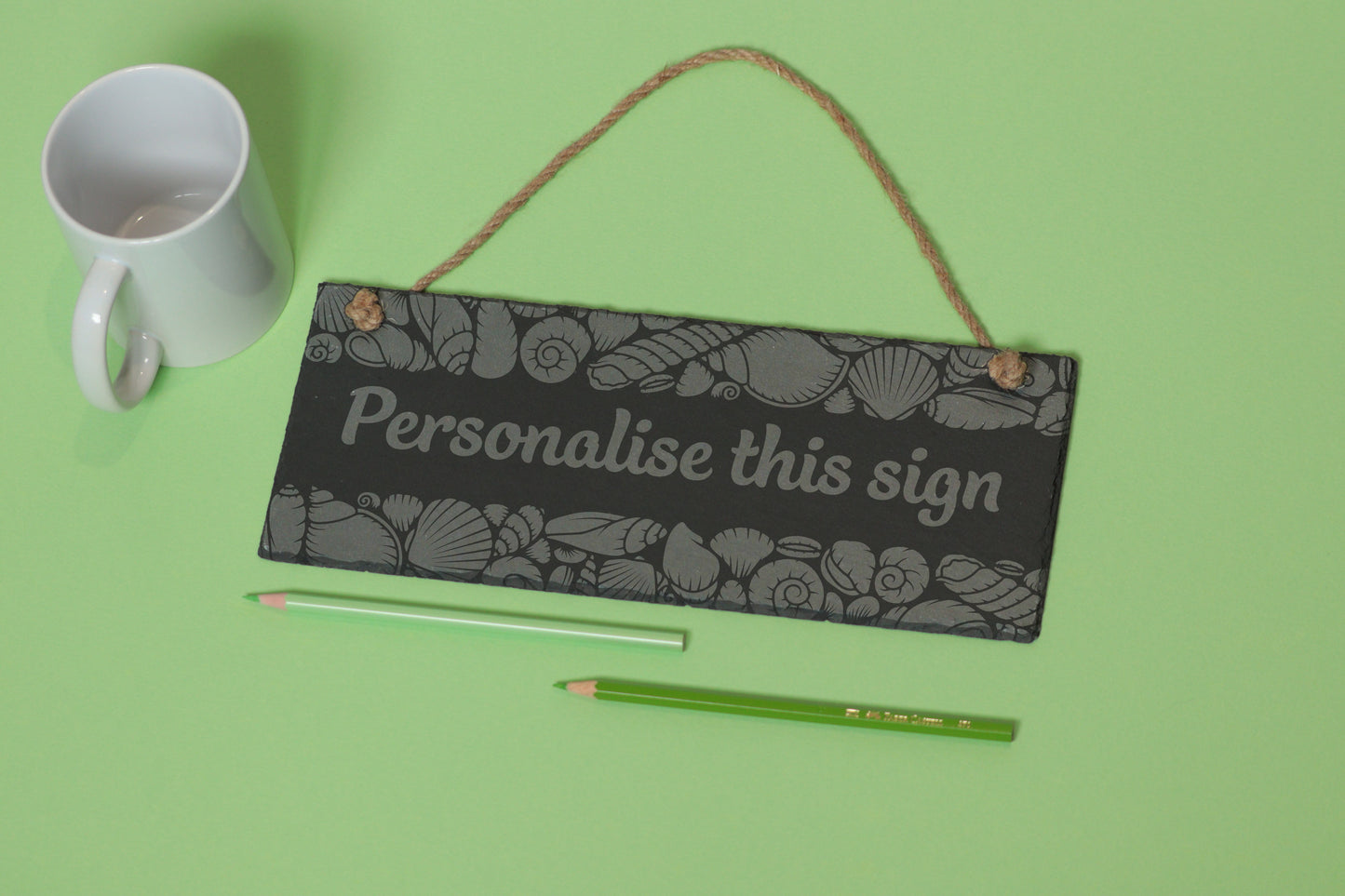 Flat-lay of a customizable slate sign with sea shell engraving and 'Personalise this sign' text, next to a white mug and two coloured pencils on a light green background.