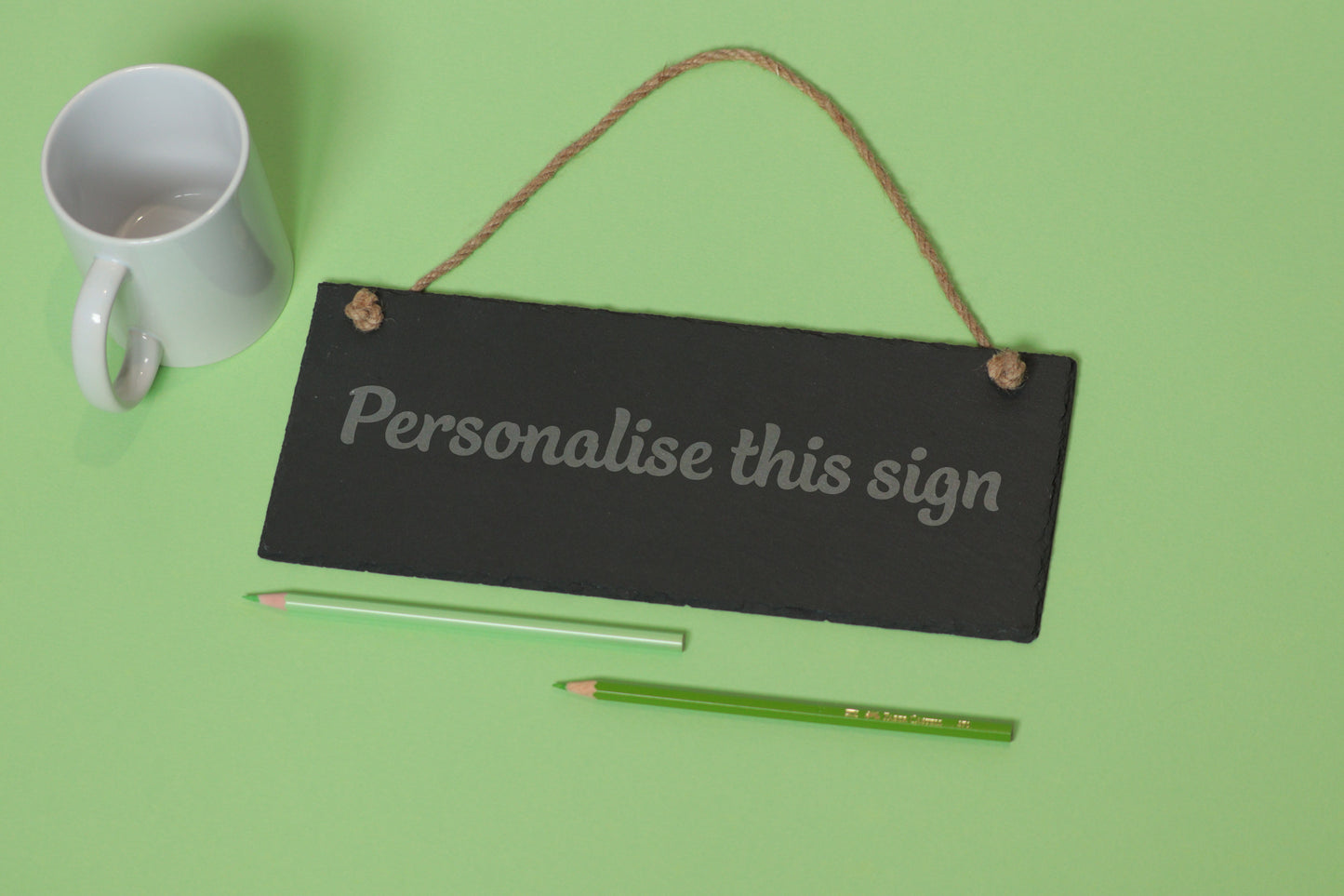 Flat-lay of a customizable slate sign with 'Personalise this sign' text, next to a white mug and two coloured pencils on a light green background.