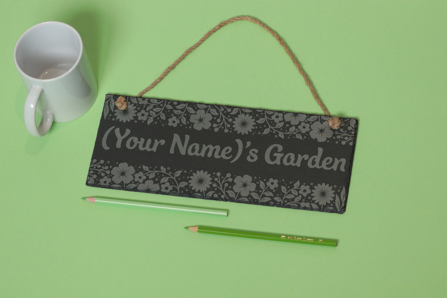 Flat-lay of a customizable slate sign with floral engraving and '(Your Name)'s Garden' text, next to a white mug and two coloured pencils on a light green background.