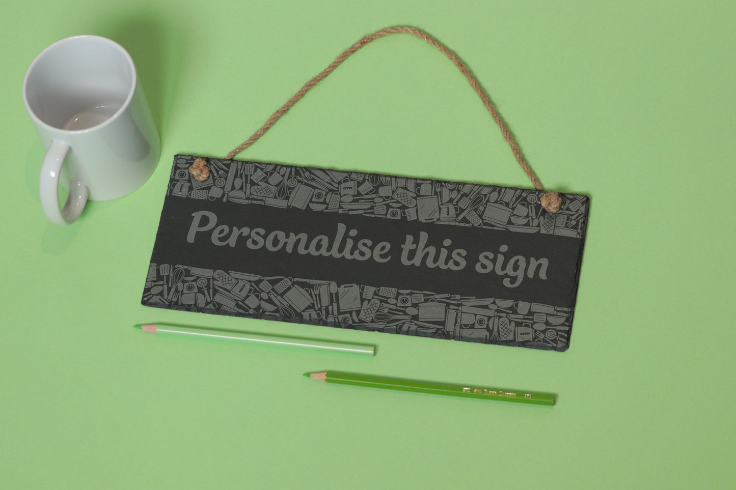 Flat-lay of a customizable slate sign with kitchen utensil engraving and 'Personalise this sign' text, next to a white mug and two coloured pencils on a light green background.