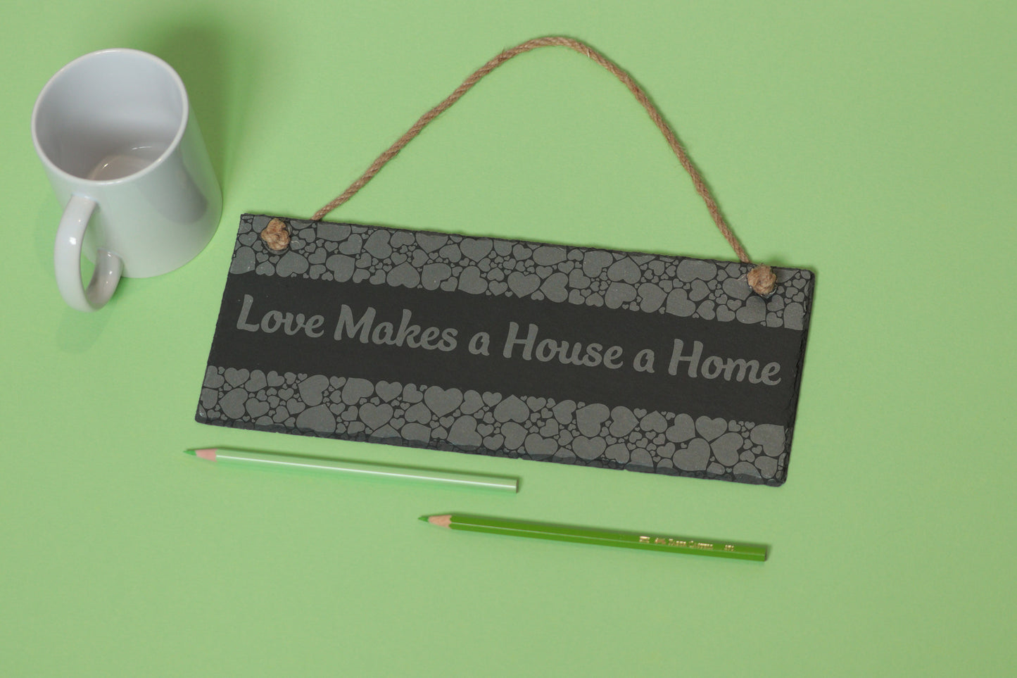 Flat-lay of a customizable slate sign with heart engraving and 'Love Makes a House a Home' text, next to a white mug and two coloured pencils on a light green background.
