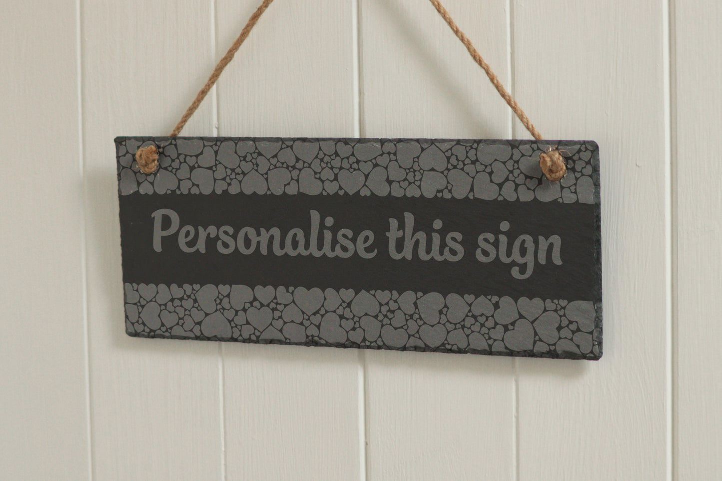 Customizable engraved slate sign with heart border, hanging on rope, with the text 'Personalise this sign', displayed against a white wooden background.
