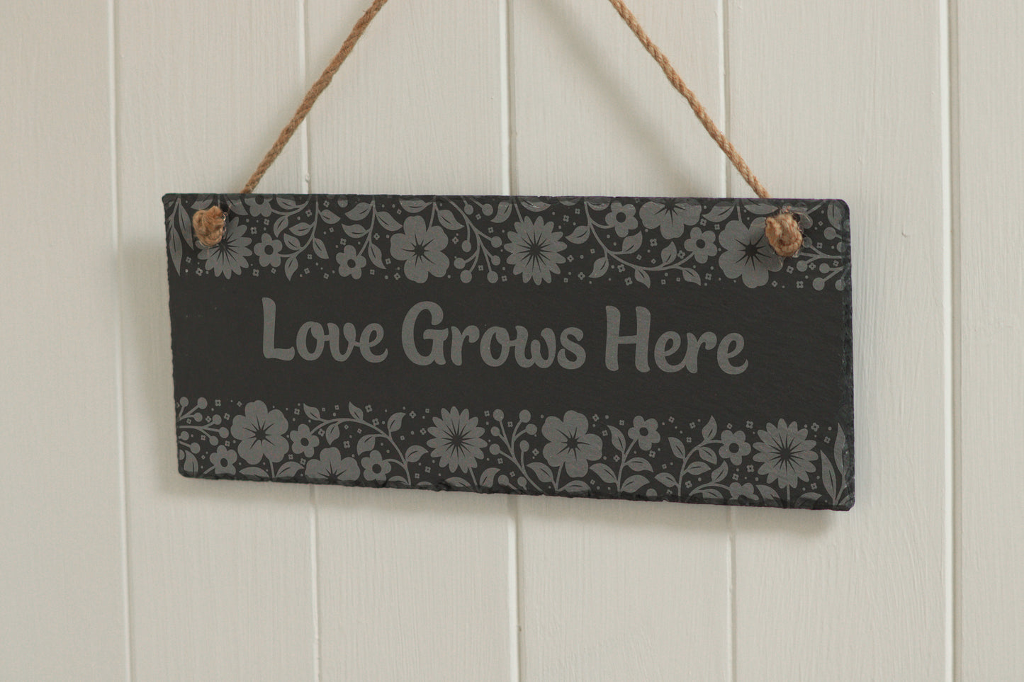 Customizable engraved slate sign with floral border, hanging on rope, with the text 'Love Grows Here', displayed against a white wooden background.