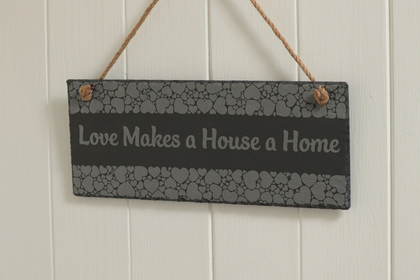 Customizable engraved slate sign with heart border, hanging on rope, with the text 'Love Makes a House a Home', displayed against a white wooden background.