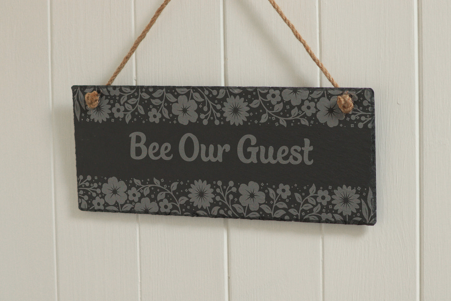Customizable engraved slate sign with floral border, hanging on rope, with the text 'Bee Our Guest', displayed against a white wooden background.