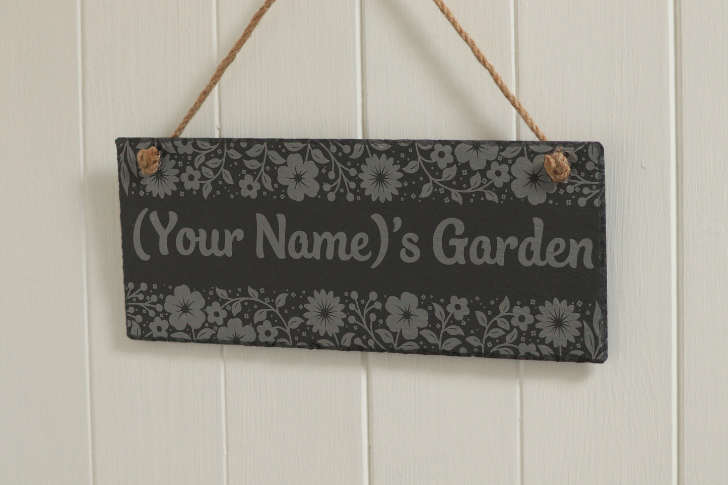 Customizable engraved slate sign with floral border, hanging on rope, with the text '(Your Name)'s Garden', displayed against a white wooden background.