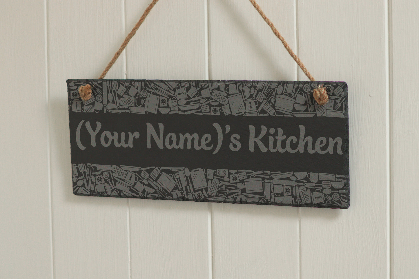 Customizable engraved slate sign with kitchen utensil border, hanging on rope, with the text '(Your Name)'s Kitchen', displayed against a white wooden background.