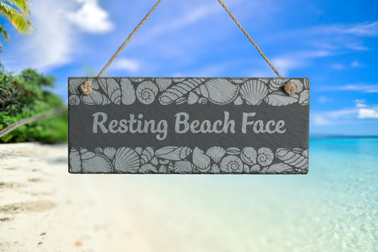 Customizable slate sign with engraved sea shell border and the text 'Resting Beach Face', hanging in a beach setting.