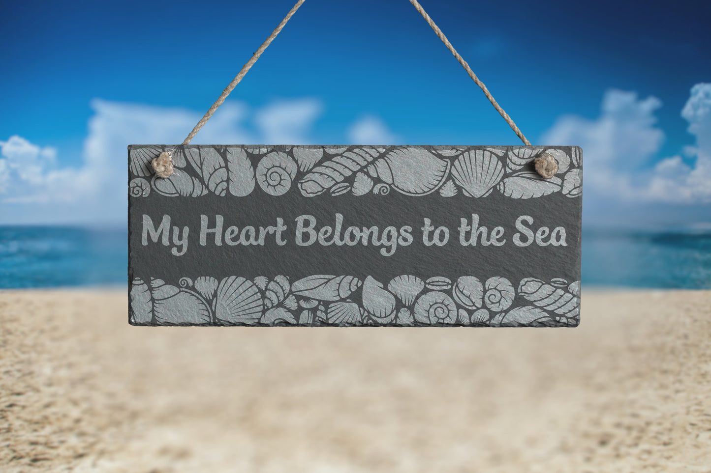 Customizable slate sign with engraved sea shell border and the text 'My Heart Belongs to the Sea', hanging in a beach setting.