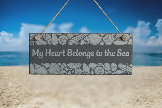 Customizable slate sign with engraved sea shell border and the text 'My Heart Belongs to the Sea', hanging in a beach setting.
