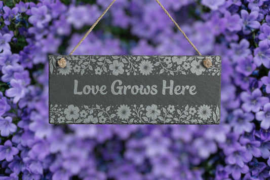 Customizable slate sign with engraved floral border and the text 'Love Grows Here', hanging in a garden setting.