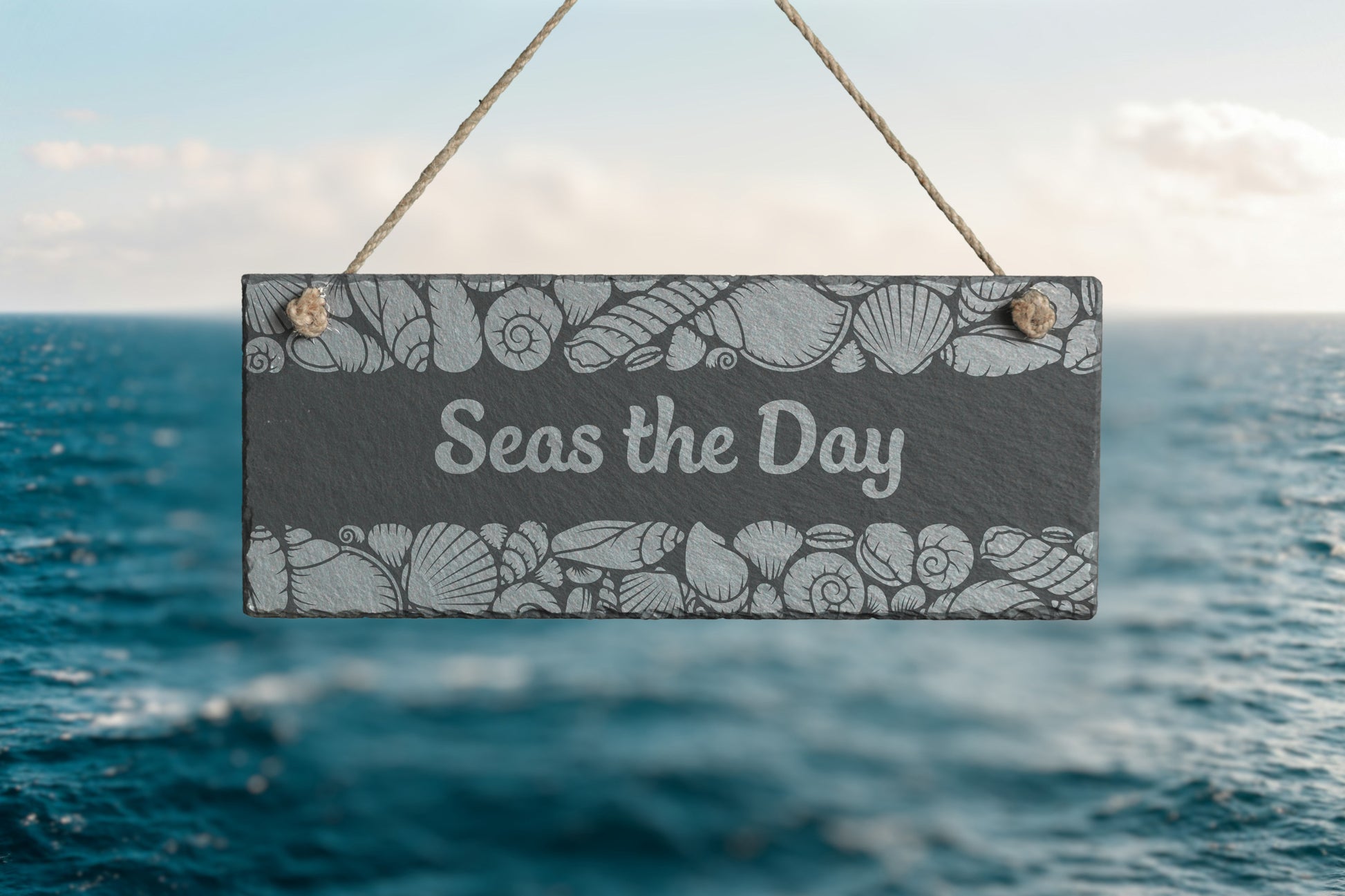 Customizable slate sign with engraved sea shell border and the text 'Seas the Day', hanging in a beach setting.