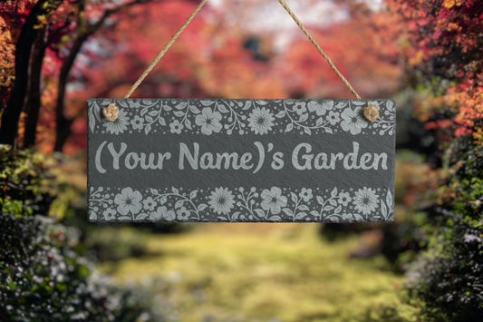 Decorative garden slate sign with floral patterns and 'Your Name's Garden' text, set against a blurred autumnal garden background.
