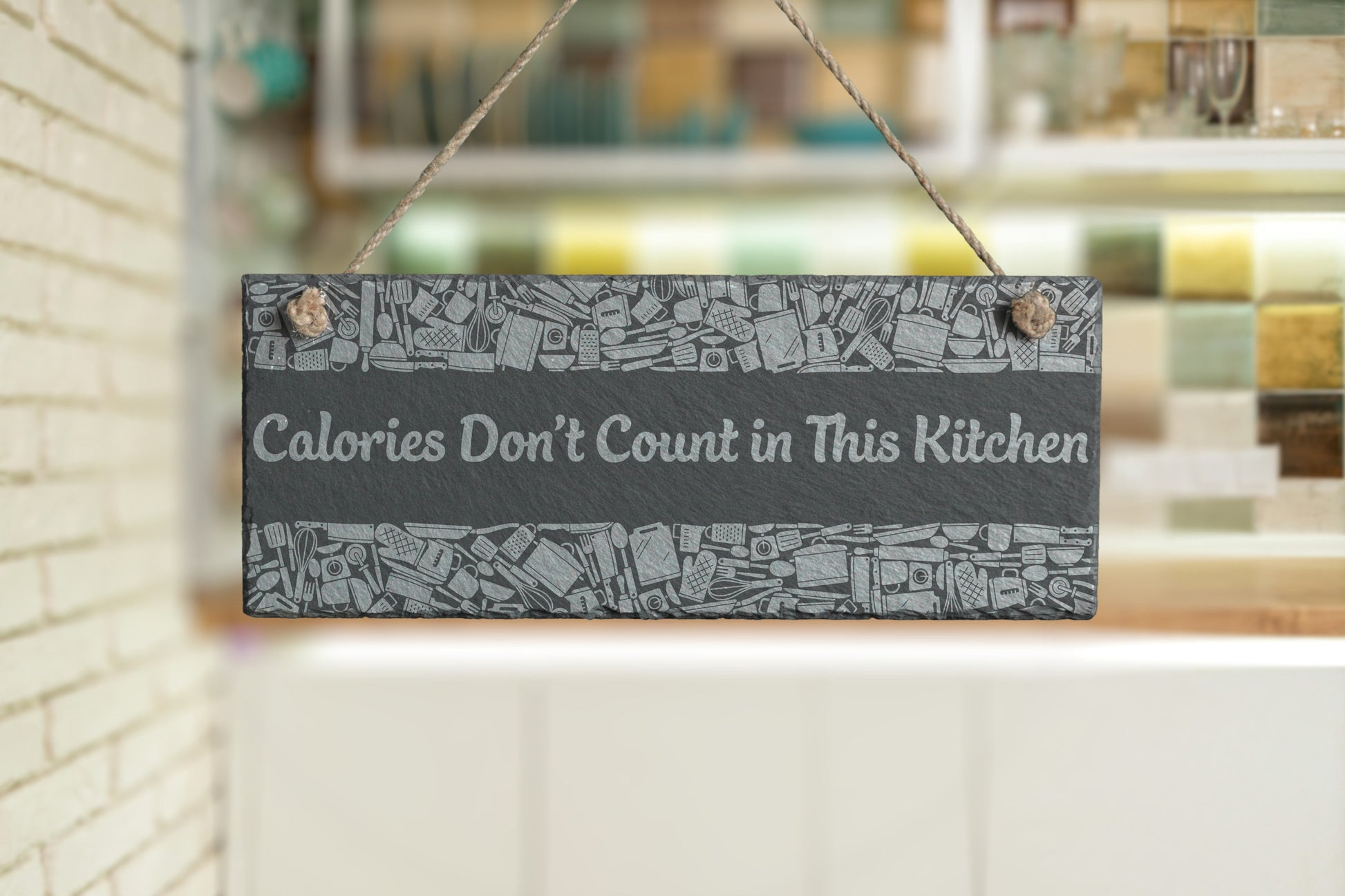 Customizable slate sign with engraved kitchen utensil border and the text 'Calories Don't Count in This Kitchen', hanging in a modern kitchen setting.