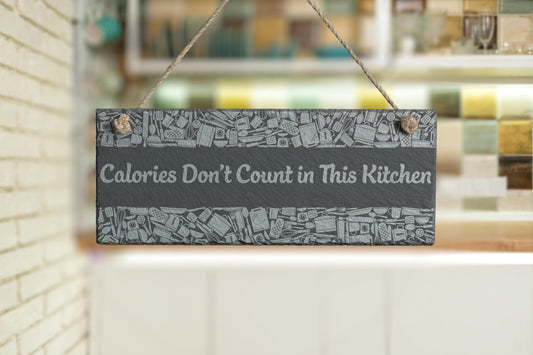 Customizable slate sign with engraved kitchen utensil border and the text 'Calories Don't Count in This Kitchen', hanging in a modern kitchen setting.