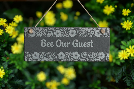 Customizable slate sign with engraved floral border and the text 'Bee Our Guest', hanging in a garden setting.