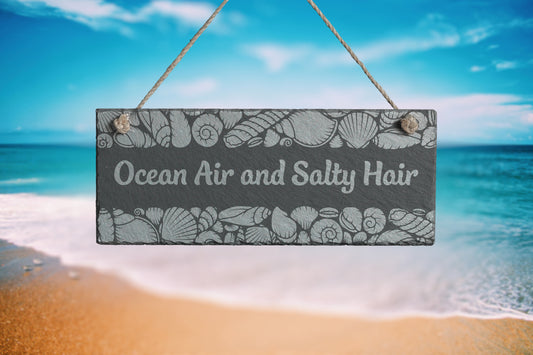 Customizable slate sign with engraved sea shell border and the text 'Ocean Air and Salty Hair', hanging in a beach setting.