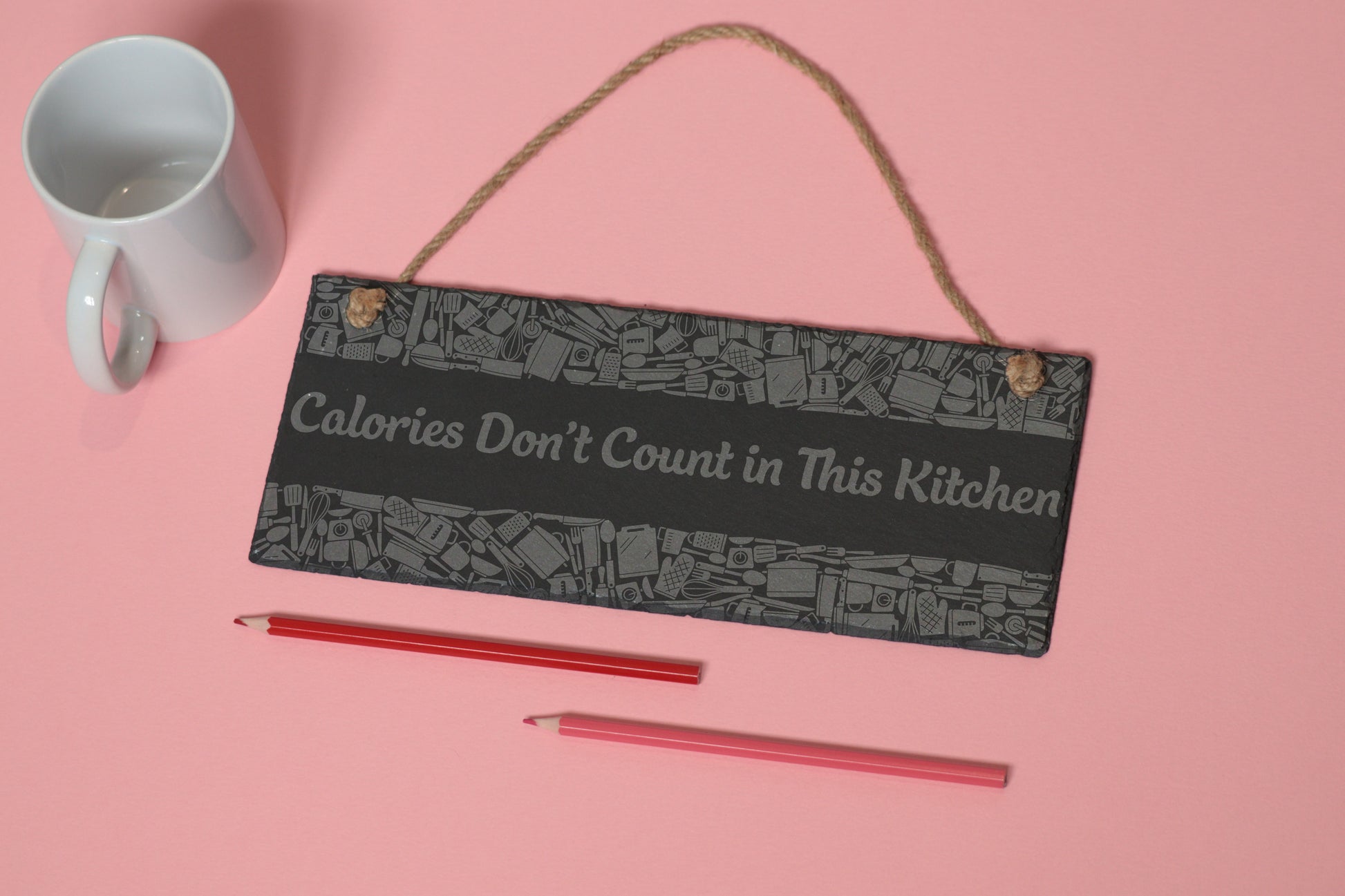 Flat-lay of a customizable slate sign with kitchen utensils engraving and 'Calories Don't Count in This Kitchen' text, next to a white mug and two coloured pencils on a light pink background.