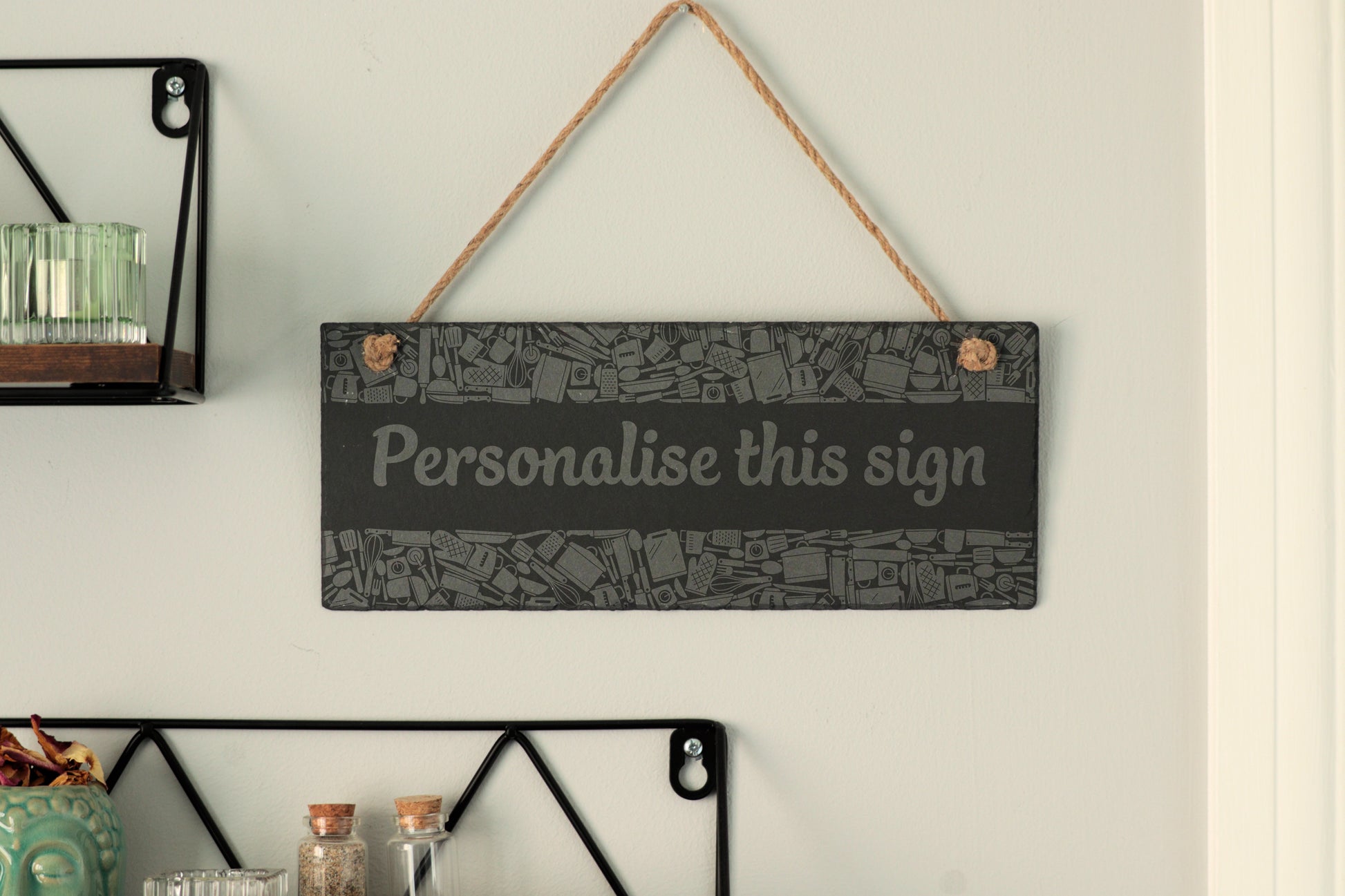 Customizable slate sign with kitchen utensil border and the text 'Personalise this sign', hanging on a wall near shelves.