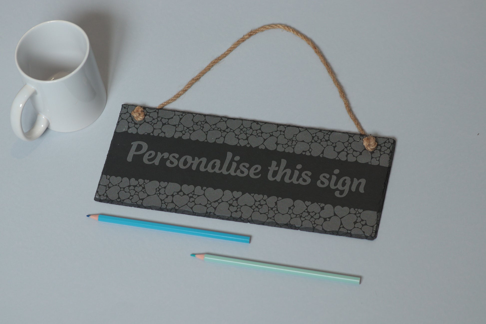 Flat-lay of a customizable slate sign with heart engraving and 'Personalise this sign' text, next to a white mug and two coloured pencils on a light blue background.