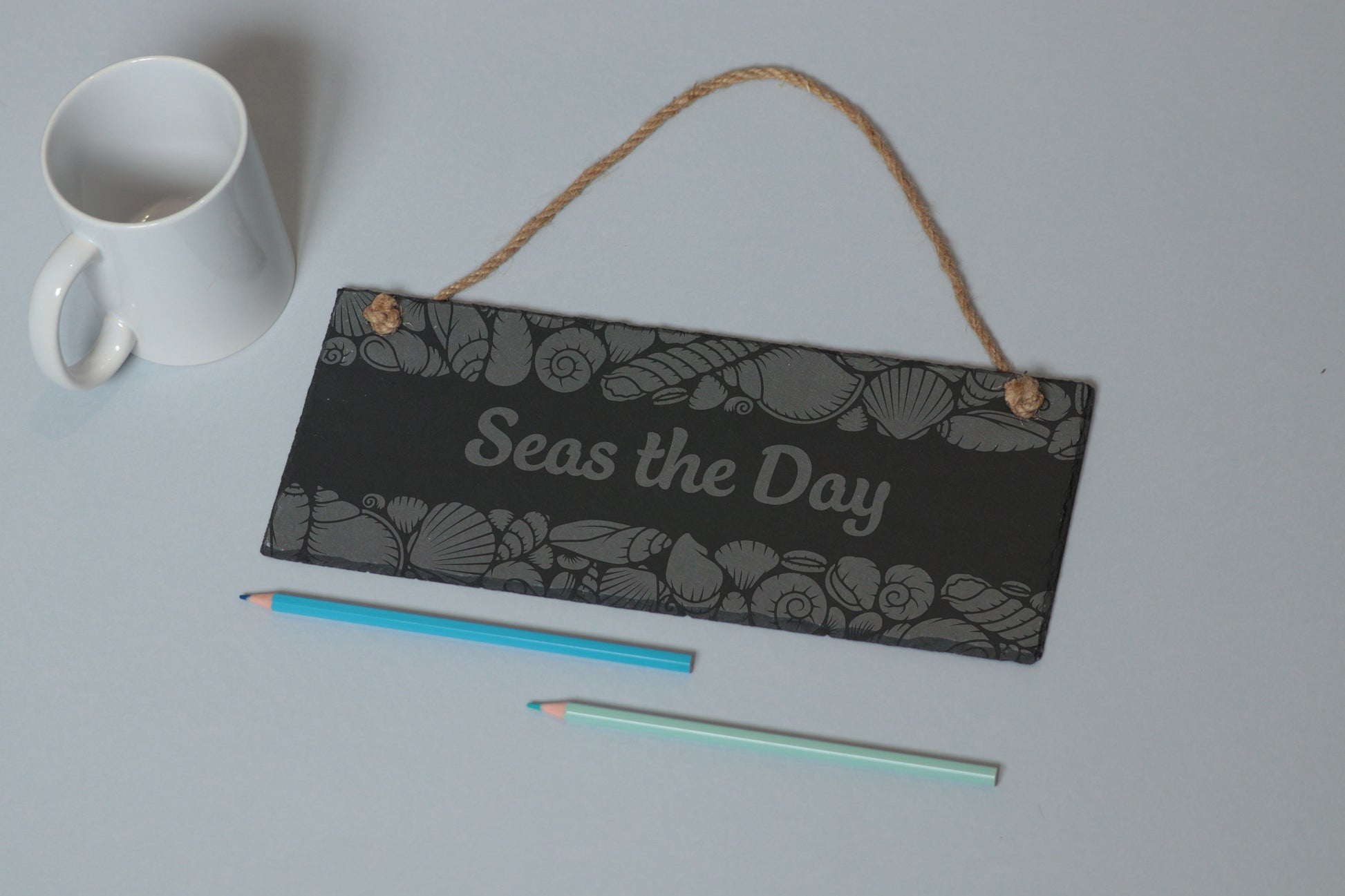 Flat-lay of a customizable slate sign with sea shell engraving and 'Seas the Day' text, next to a white mug and two coloured pencils on a light blue background.