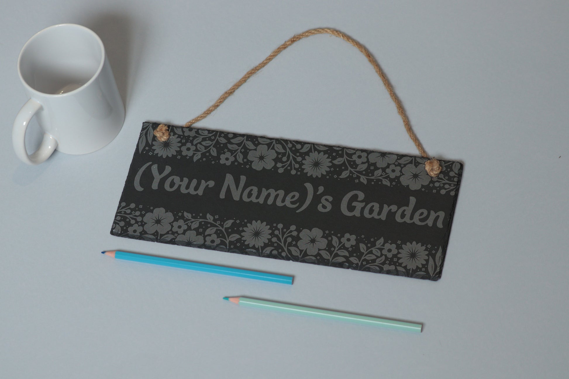 Flat-lay of a customizable slate sign with floral utensil engraving and '(Your Name)'s Garden' text, next to a white mug and two coloured pencils on a light blue background.