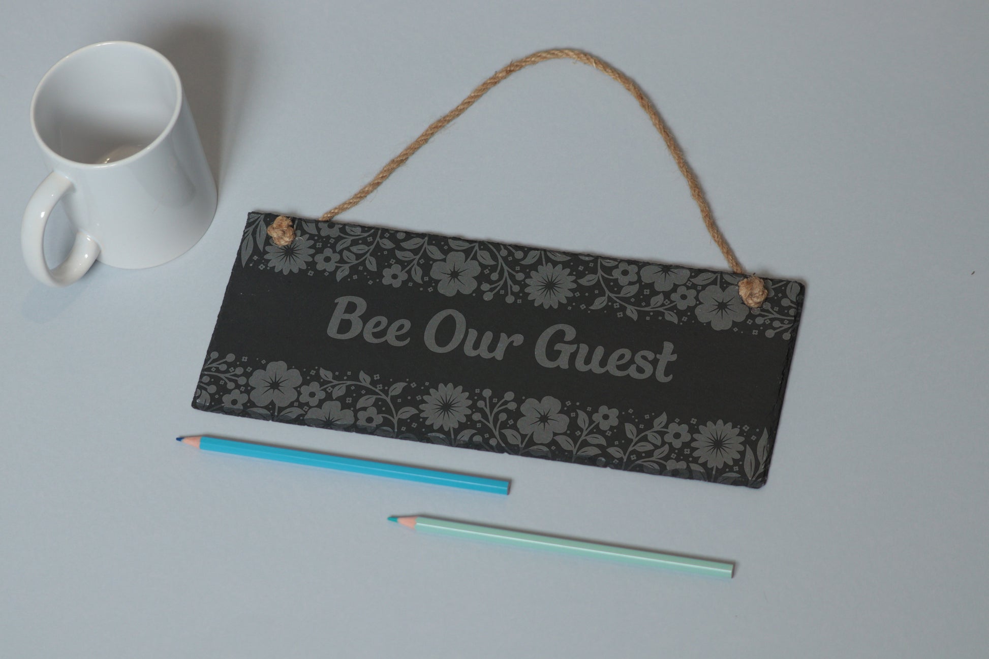 Flat-lay of a customizable slate sign with floral engraving and 'Bee Our Guest' text, next to a white mug and two coloured pencils on a light blue background.