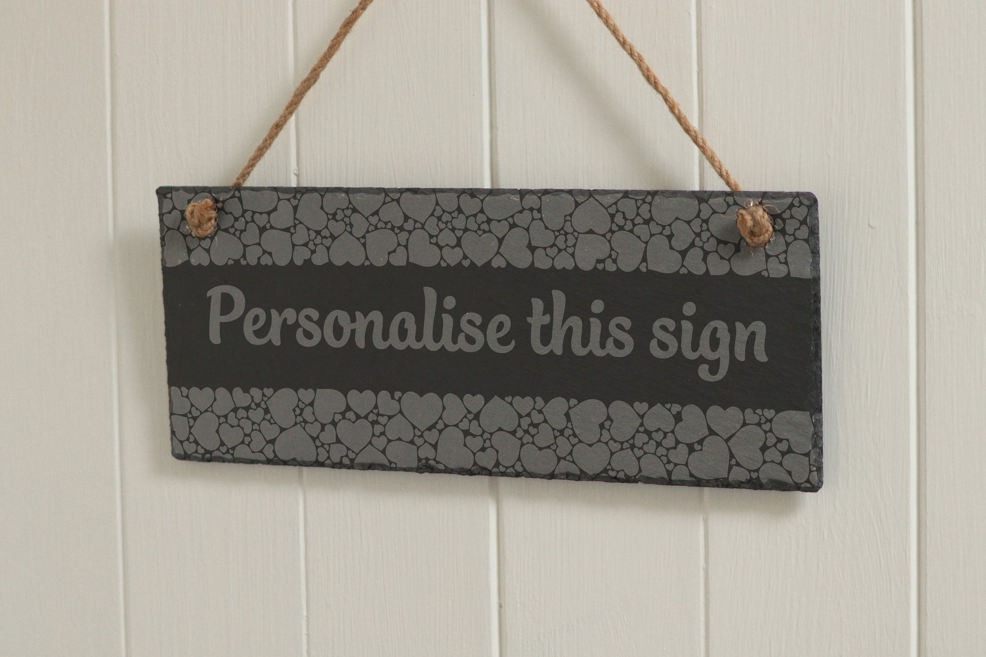 Customizable engraved slate sign with heart border, hanging on rope, with the text 'Personalise this sign', displayed against a white wooden background.