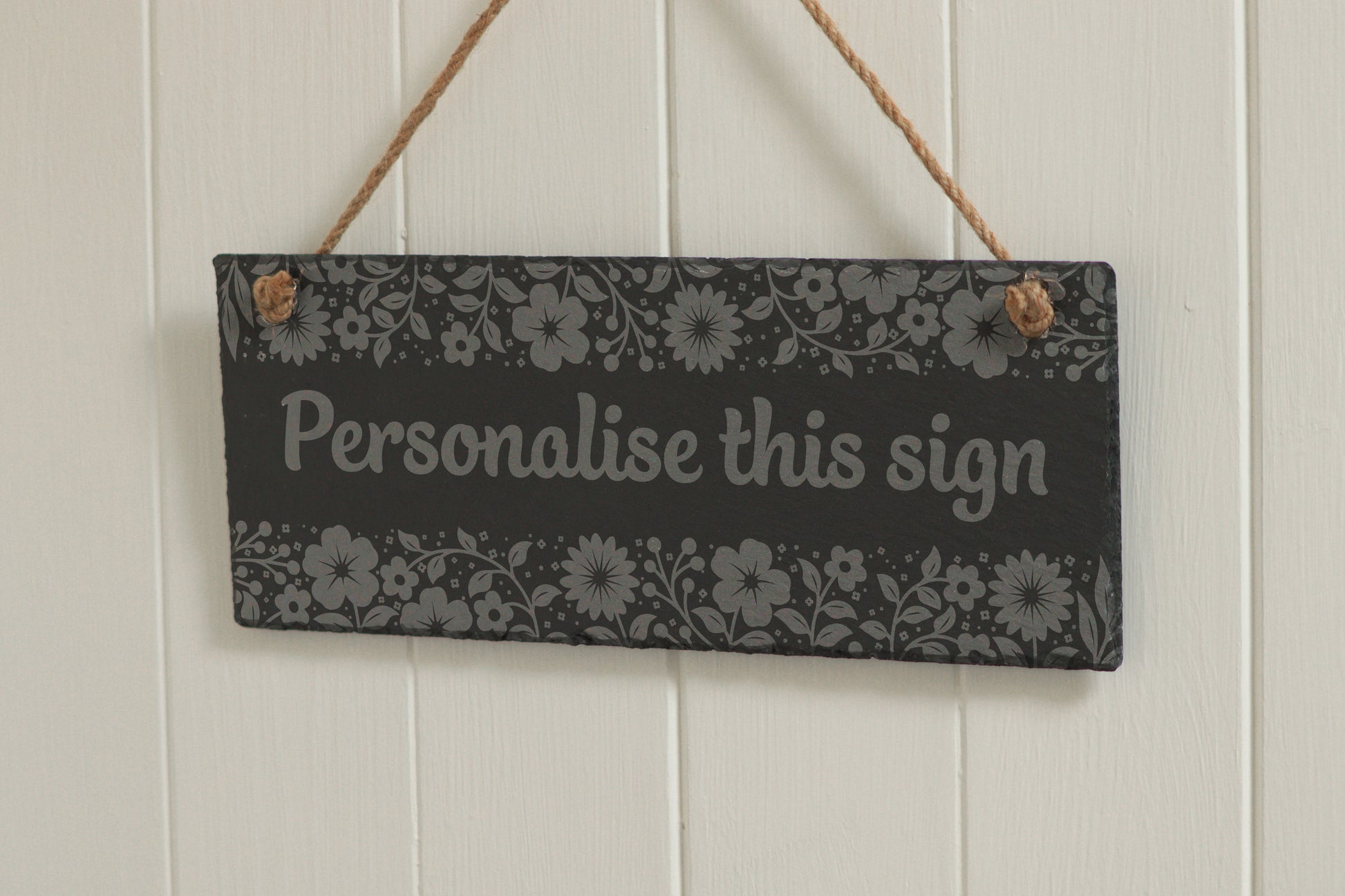Customizable engraved slate sign with floral border, hanging on rope, with the text 'Personalise this sign', displayed against a white wooden background.