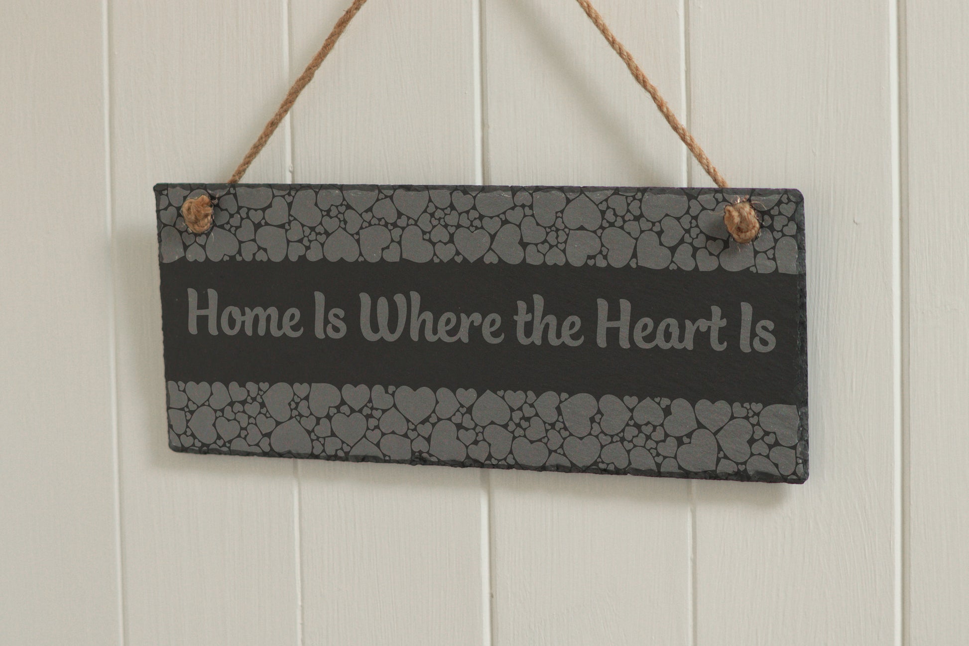 Customizable engraved slate sign with heart border, hanging on rope, with the text 'Home is Where the Heart Is', displayed against a white wooden background.